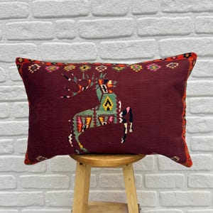 May include: A burgundy throw pillow with a colourful geometric design of a deer. The pillow has a fringe border and is sitting on a wooden stool.