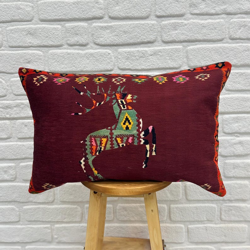 May include: A burgundy throw pillow with a colourful geometric design of a deer. The pillow has a fringe border and is sitting on a wooden stool.