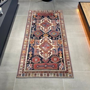 Op de afbeelding: Een traditioneel tapijt met een geometrisch patroon in tinten rood, blauw en wit. Het tapijt heeft een donkerblauwe achtergrond met een centrale ruitvorm omgeven door kleinere geometrische vormen.