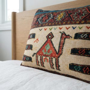 May include: Rectangular beige pillow with a camel design in red, orange, and green. The pillow has a patterned border in red, black, and blue. The pillow is placed on a white bed.