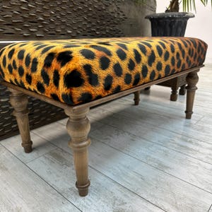 May include: A rectangular bench with a leopard print seat cushion and wooden legs. The cushion features a pattern of black spots on a tan and orange background. The legs are light brown and have a turned design.