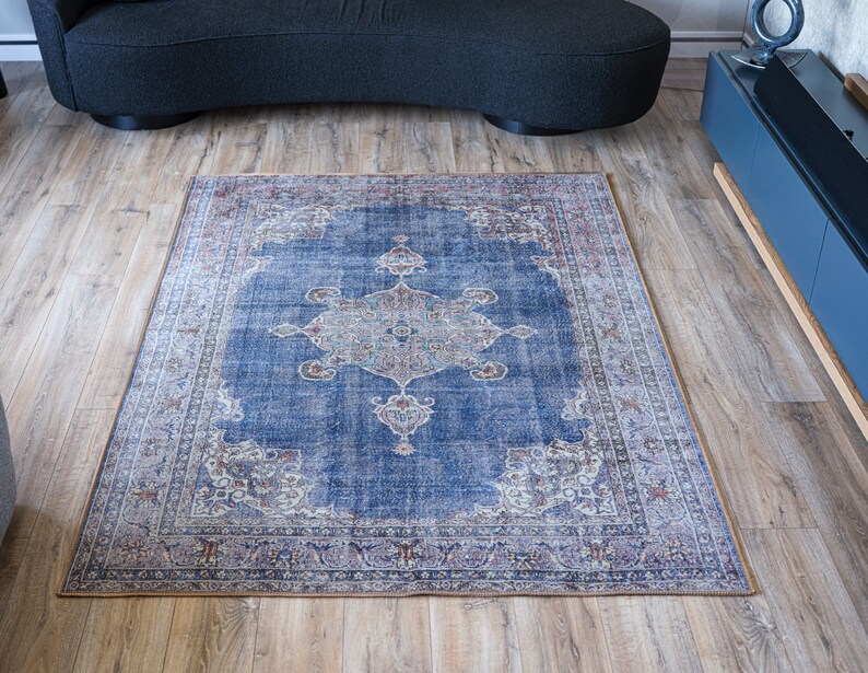 Op de afbeelding: Een blauw en cr&egrave;mekleurig tapijt met een bloemmotief in het midden. Het tapijt heeft een verweerde uitstraling en ligt op een houten vloer.