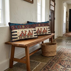 May include: Two rectangular throw pillows with a camel design rest on a wooden bench. The pillows have a cream background with a camel motif in red, orange, and blue. The bench is made of light brown wood, and a woven basket sits below.
