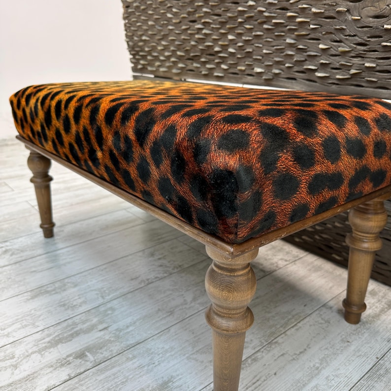 May include: A rectangular bench with a leopard print seat cushion. The cushion features a pattern of black spots on an orange background. The bench has four wooden legs and a wooden frame.