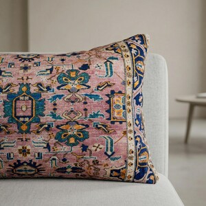 May include: A rectangular decorative pillow with a vintage rug design. The pillow features a dusty rose background with intricate patterns in blue, yellow, and teal. A navy blue border frames the design, adding a touch of elegance.