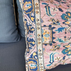 May include: Close-up of a decorative pillow with a vintage rug design. The pillow features a dusty rose background with intricate patterns in navy blue, gold, and teal. The pillow is placed on a dark gray surface.