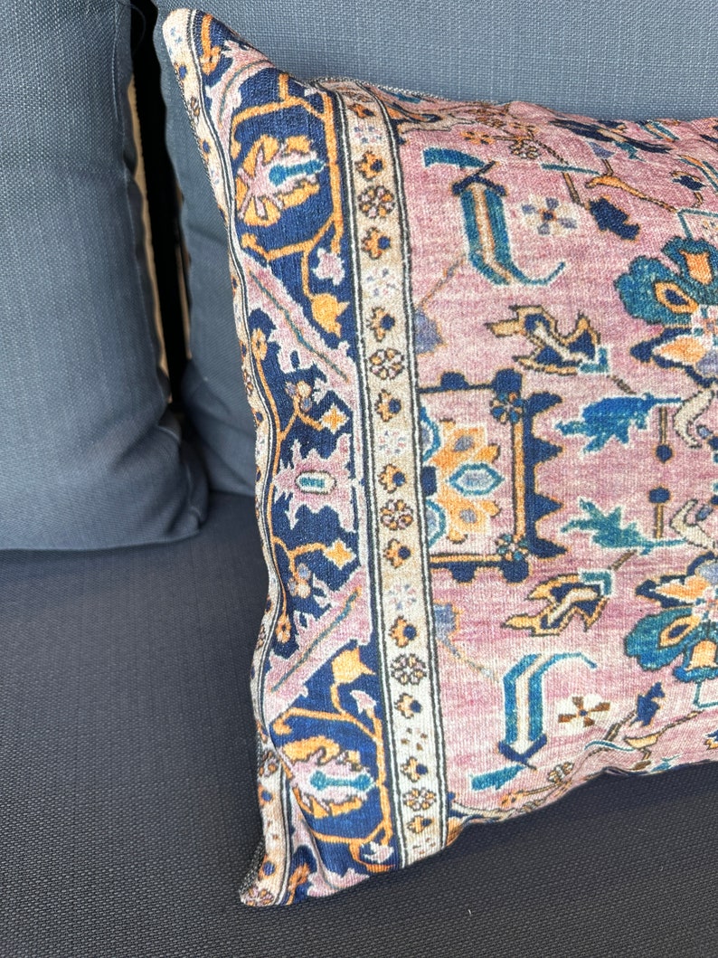 May include: Close-up of a decorative pillow with a vintage rug design. The pillow features a dusty rose background with intricate patterns in navy blue, gold, and teal. The pillow is placed on a dark gray surface.