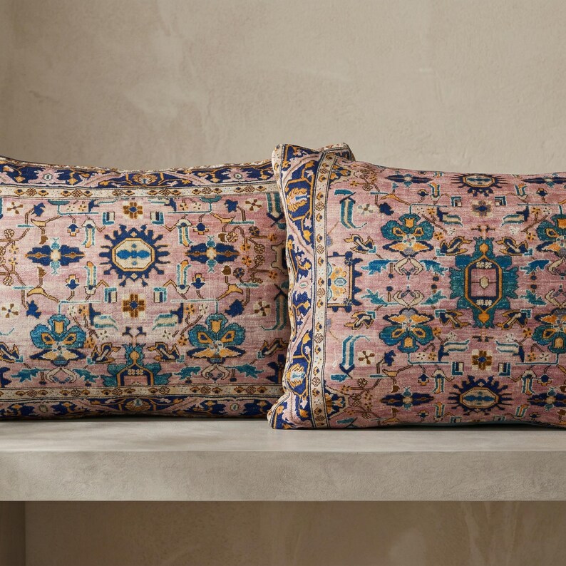 May include: Two decorative throw pillows with a vintage rug design. The pillows feature a dusty rose background with intricate patterns in shades of blue, gold, and teal. The design includes geometric shapes and floral motifs, creating a classic, elegant look.