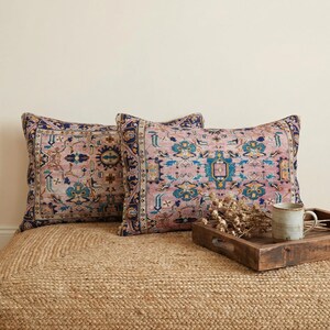 May include: Two decorative throw pillows with a vintage rug design in shades of pink, blue, and gold. The pillows are square and rest on a woven jute rug. A wooden tray with a mug and dried flowers sits nearby.
