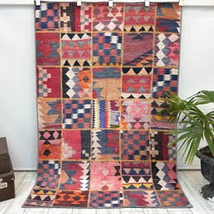 May include: A large, rectangular patchwork rug with a vibrant mix of colors and geometric patterns. The rug features squares of red, blue, orange, and beige, creating a textured and visually appealing design. It is hanging against a white wall.