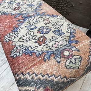 May include: A close-up of a brown and beige patterned ottoman with a dark blue and red geometric design. The ottoman is upholstered in a fabric that resembles a rug.