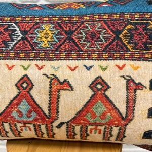 May include: Decorative pillow with a beige background and a camel design in red, orange, and green. The pillow features a blue and red geometric border. The camel design is repeated across the pillow's surface.
