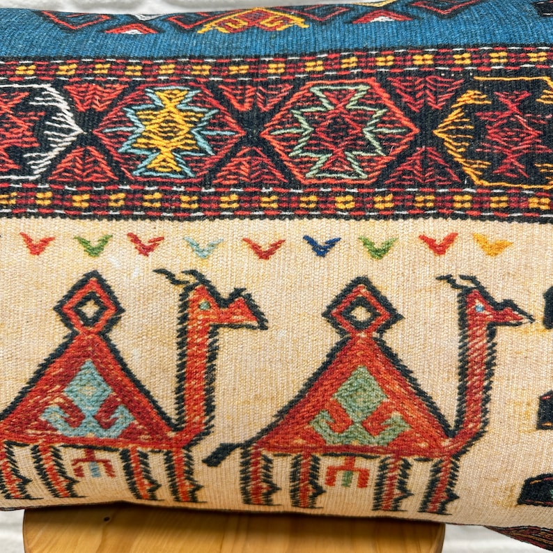 May include: Decorative pillow with a beige background and a camel design in red, orange, and green. The pillow features a blue and red geometric border. The camel design is repeated across the pillow's surface.