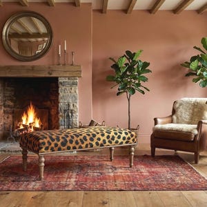 May include: A cozy living room scene with a fireplace, a leopard-print bench, and a leather armchair. The room features a rug, plants, and a round mirror above the fireplace. The walls are painted in a warm, earthy tone.