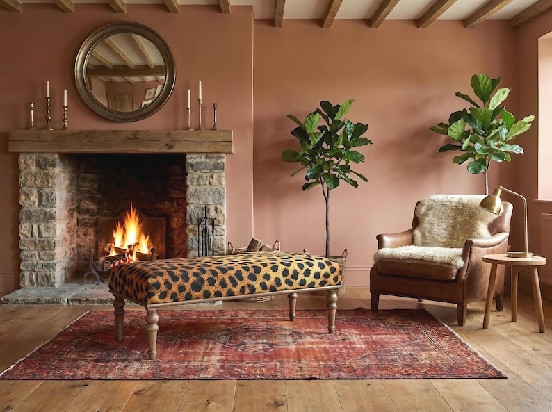 May include: A cozy living room scene with a fireplace, a leopard-print bench, and a leather armchair. The room features a rug, plants, and a round mirror above the fireplace. The walls are painted in a warm, earthy tone.
