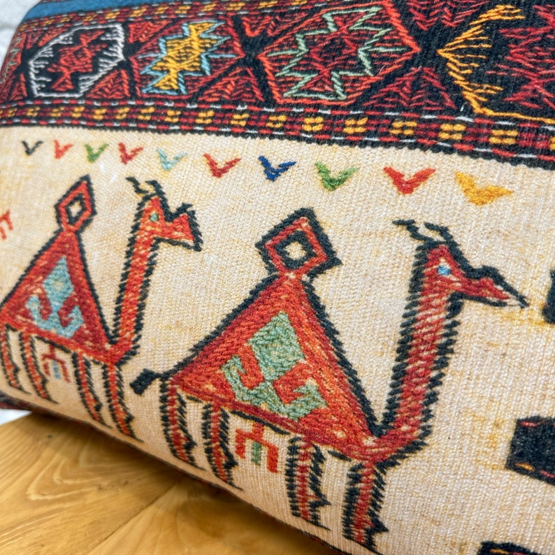 May include: Close-up of a rectangular pillow with a beige background and a camel design. The camels are red with blue and green accents. The top border features a geometric pattern in red, yellow, and black. The pillow has a blue border.