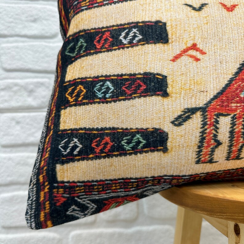 May include: Close-up of a decorative pillow with a beige background and a detailed pattern. The pattern includes black, red, and green geometric shapes and lines. The pillow is placed on a wooden stool against a white brick wall.