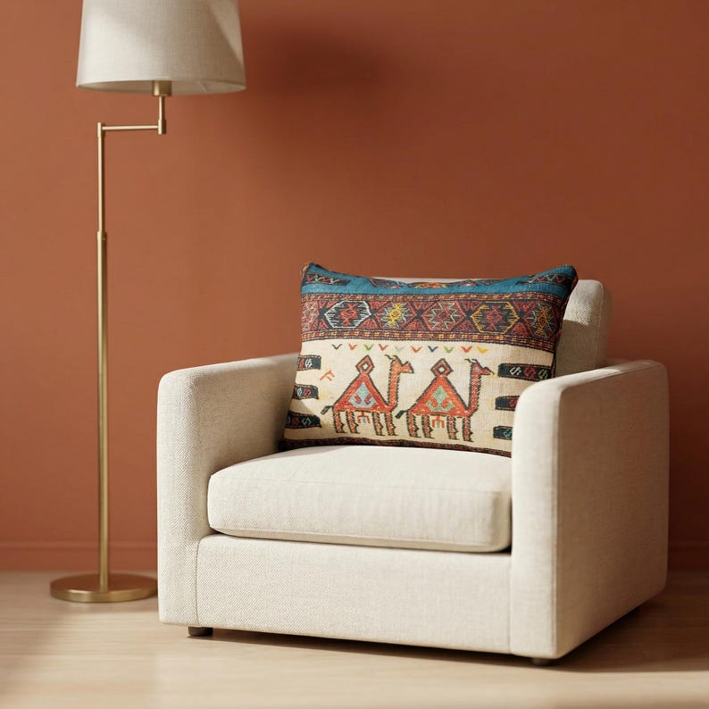 May include: A cream-colored armchair with a decorative pillow featuring a camel design. The pillow has a blue border and a cream background with red, orange, and yellow accents. A gold floor lamp stands to the left.