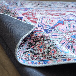K&ouml;nnte beinhalten: Nahaufnahme eines bunten Teppichs mit einem rosa, blauen und wei&szlig;en Muster. Der Teppich ist umgeklappt und zeigt eine dunkelgraue R&uuml;ckseite.