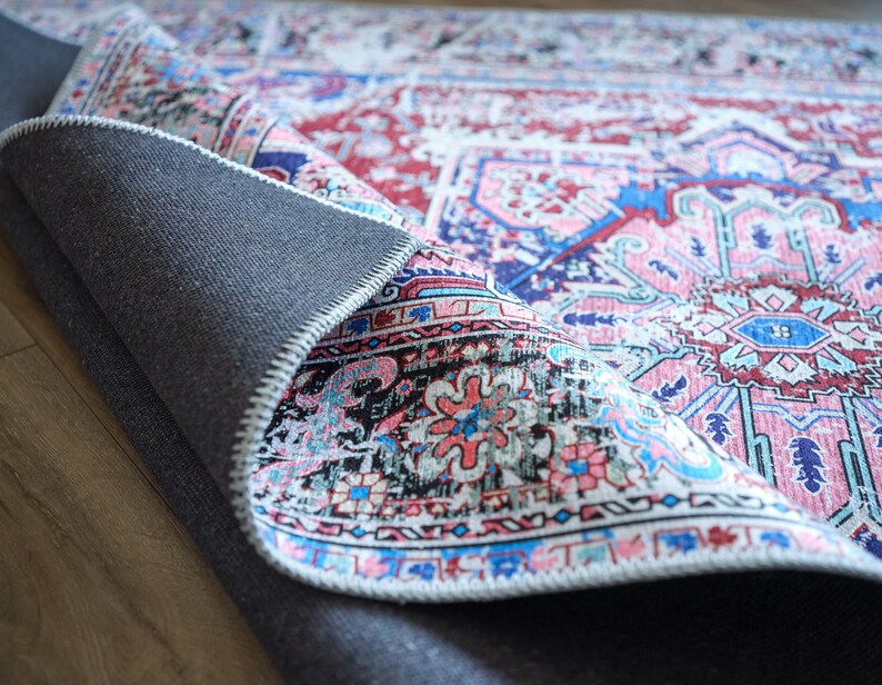 K&ouml;nnte beinhalten: Nahaufnahme eines bunten Teppichs mit einem rosa, blauen und wei&szlig;en Muster. Der Teppich ist umgeklappt und zeigt eine dunkelgraue R&uuml;ckseite.