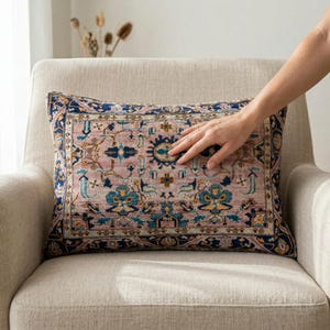 May include: Rectangular decorative pillow with a vintage rug design. The pillow features a pink center with blue and teal floral patterns and a navy blue border. The pillow is placed on a beige armchair.