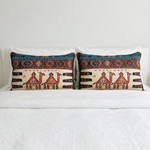 May include: Two rectangular throw pillows with a cream-colored background and a camel design. The pillows have a teal and patterned border. The camel design is in shades of red, orange, and brown. The pillows are on a white bed.