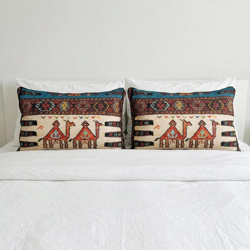 May include: Two rectangular throw pillows with a cream-colored background and a camel design. The pillows have a teal and patterned border. The camel design is in shades of red, orange, and brown. The pillows are on a white bed.