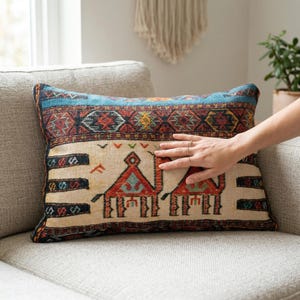 May include: Rectangular decorative pillow with a patterned design. The pillow features a blue border with geometric patterns in red, yellow, and black. The center has a beige background with two camel figures and additional geometric designs.