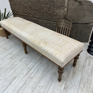 May include: A long, upholstered bench with a cream-colored fabric featuring a gold pattern. The bench has dark wooden legs and a rectangular shape. The design is classic and would fit well in a living room or entryway.