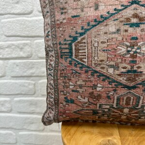 May include: A pink and brown patterned throw pillow with a teal border. The pillow is sitting on a wooden chair.