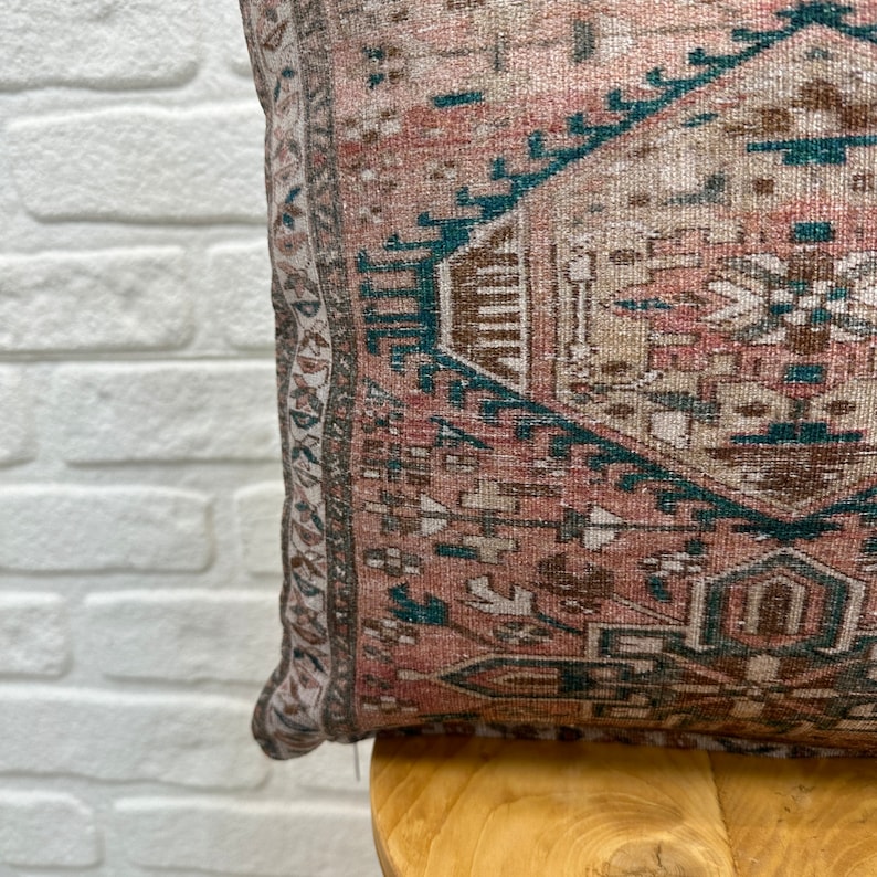 May include: A pink and brown patterned throw pillow with a teal border. The pillow is sitting on a wooden chair.