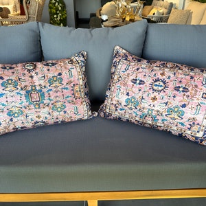 May include: Two rectangular throw pillows with a vintage rug design. The pillows feature a pink background with a detailed pattern in shades of blue, teal, and yellow. The borders are dark blue.
