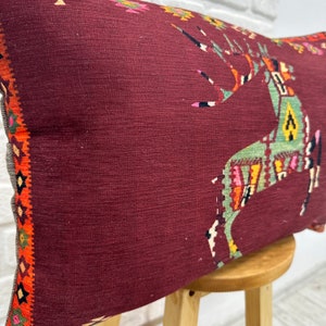 May include: A maroon throw pillow with a colourful geometric design of a deer. The pillow has a fringe border.