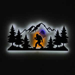 Puede incluir: Una silueta de metal negro de un Bigfoot llevando una bandera estadounidense, caminando por una cordillera con &aacute;rboles a cada lado. La silueta est&aacute; iluminada con una luz blanca.