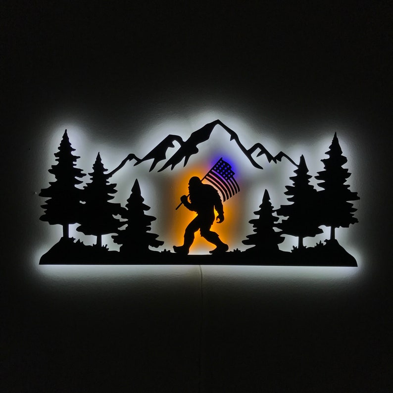 Puede incluir: Una silueta de metal negro de un Bigfoot llevando una bandera estadounidense, caminando por una cordillera con &aacute;rboles a cada lado. La silueta est&aacute; iluminada con una luz blanca.