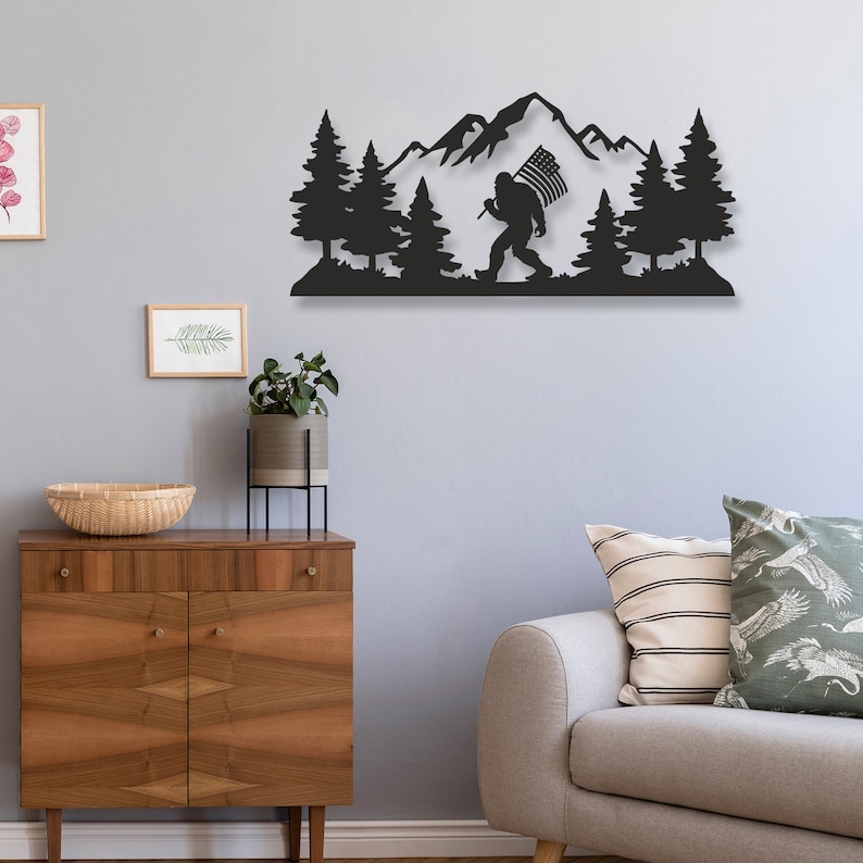 Puede incluir: Arte de pared de metal negro con una silueta de Bigfoot llevando una bandera estadounidense, caminando por un bosque con monta&ntilde;as en el fondo.