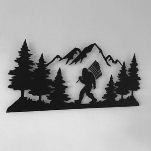 Puede incluir: Arte de pared de metal negro que representa una silueta de Bigfoot llevando una bandera estadounidense frente a una cordillera y pinos.
