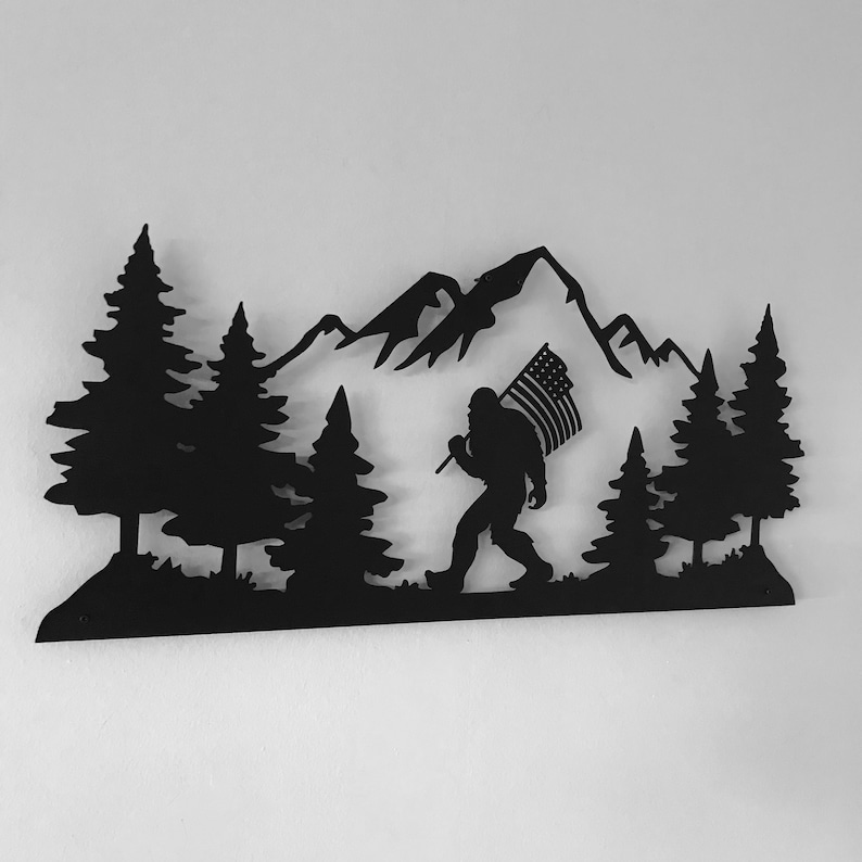 Puede incluir: Arte de pared de metal negro que representa una silueta de Bigfoot llevando una bandera estadounidense frente a una cordillera y pinos.
