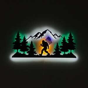 Puede incluir: Una silueta de metal de Bigfoot caminando por una cordillera con una bandera estadounidense en la mano. La silueta est&aacute; iluminada desde atr&aacute;s con una luz blanca, creando un efecto de brillo. El fondo es una habitaci&oacute;n oscura.