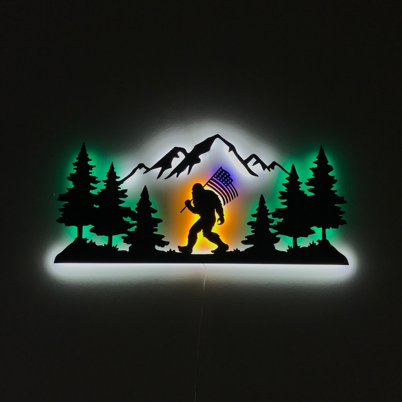 Puede incluir: Una silueta de metal de Bigfoot caminando por una cordillera con una bandera estadounidense en la mano. La silueta est&aacute; iluminada desde atr&aacute;s con una luz blanca, creando un efecto de brillo. El fondo es una habitaci&oacute;n oscura.