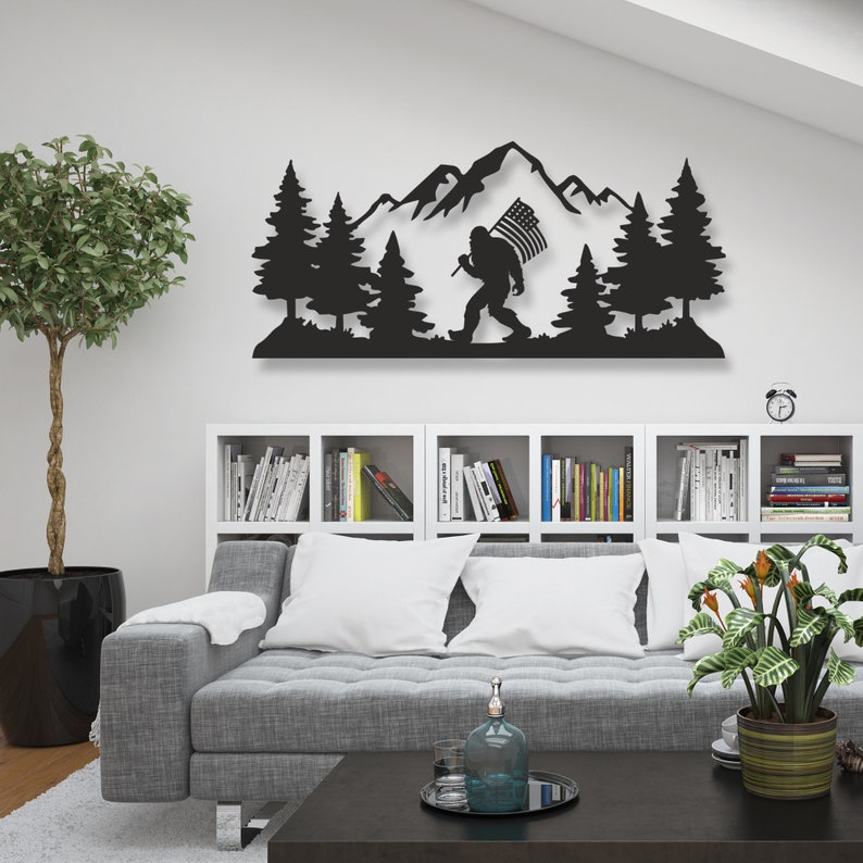 Puede incluir: Arte de pared de metal negro que representa una silueta de Bigfoot llevando una bandera estadounidense, caminando por una cordillera con pinos a cada lado.