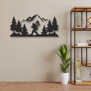Puede incluir: Arte de pared de metal negro que representa una silueta de Bigfoot caminando por un bosque con una cordillera en el fondo. El Bigfoot lleva una bandera estadounidense.