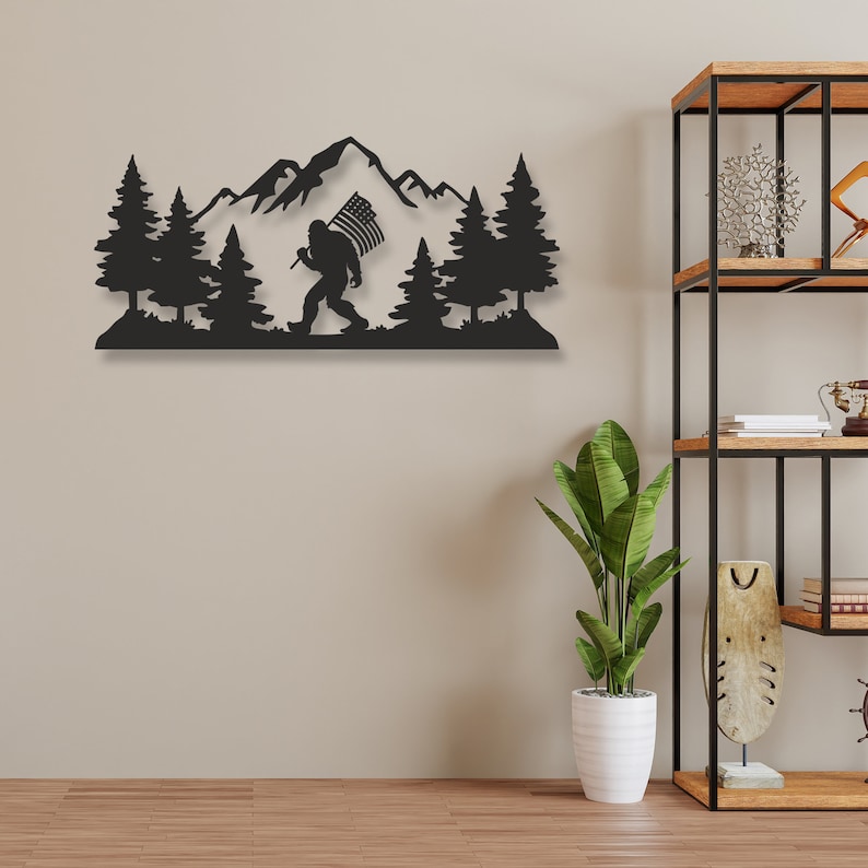Puede incluir: Arte de pared de metal negro que representa una silueta de Bigfoot caminando por un bosque con una cordillera en el fondo. El Bigfoot lleva una bandera estadounidense.