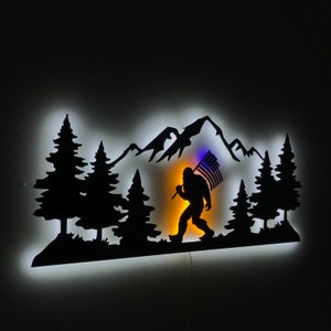 Puede incluir: Una silueta de metal negro de un Bigfoot caminando por una cordillera con una bandera estadounidense en la mano. La silueta est&aacute; iluminada desde atr&aacute;s con una luz blanca, creando un efecto de brillo.