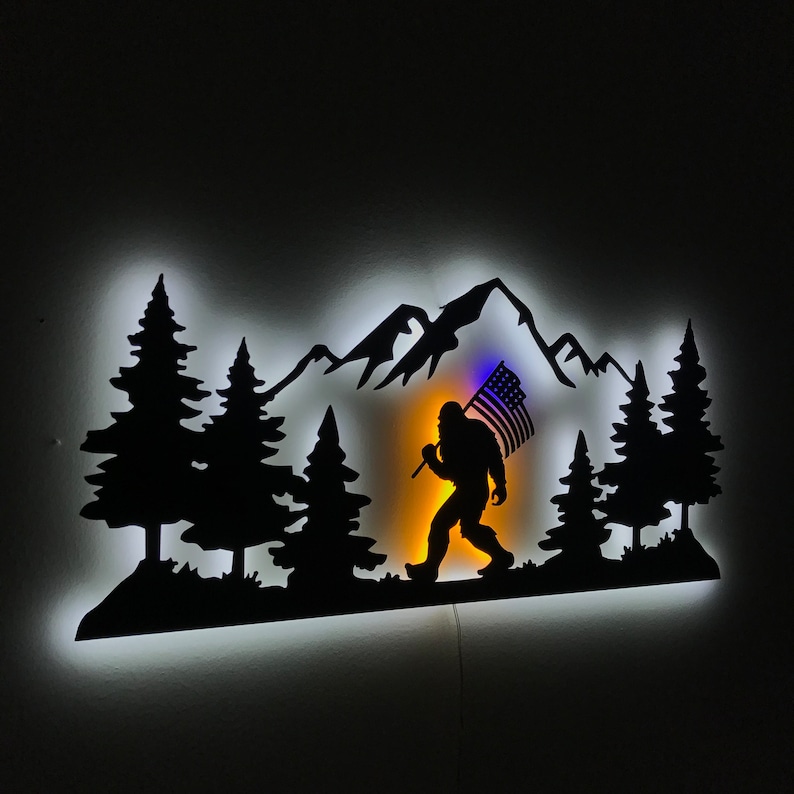 Puede incluir: Una silueta de metal negro de un Bigfoot caminando por una cordillera con una bandera estadounidense en la mano. La silueta est&aacute; iluminada desde atr&aacute;s con una luz blanca, creando un efecto de brillo.