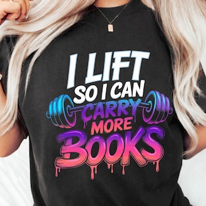 Puede incluir: Camiseta negra con el texto "I Lift So I Can Carry More Books" en blanco, azul y rosa. El diseño incluye un gráfico de barra. Una prenda de vestir informal.