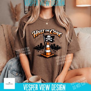 Peut inclure: T-shirt marron avec l'inscription "Hoist the Cone" et un graphisme de c&ocirc;ne de signalisation avec un drapeau t&ecirc;te de mort. Le motif comprend une silhouette de paysage urbain et est imprim&eacute; en orange, noir et blanc.