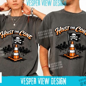 Peut inclure: T-shirts gris fonc&eacute; avec l'inscription "Hoist the Cone" en orange et blanc. Le motif repr&eacute;sente un c&ocirc;ne de signalisation, un drapeau avec une t&ecirc;te de mort et des os crois&eacute;s, et un paysage urbain. La marque "Vesper View Design" est &eacute;galement visible.
