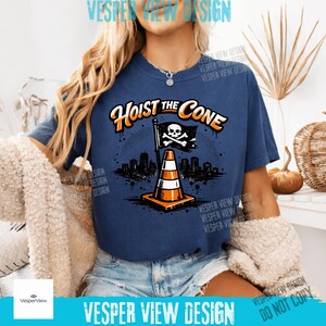 Peut inclure: T-shirt bleu marine avec l'inscription "Hoist the Cone" en orange et blanc. Le motif repr&eacute;sente un c&ocirc;ne de signalisation avec un drapeau t&ecirc;te de mort et une ligne d'horizon urbaine. C'est un v&ecirc;tement d&eacute;contract&eacute;.