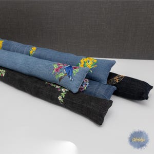 May include: A collection of decorative door draft stoppers. The stoppers are made of denim and dark gray fabric, with embroidered floral and bird designs in various colors. The stoppers are cylindrical and rest on a white surface.
